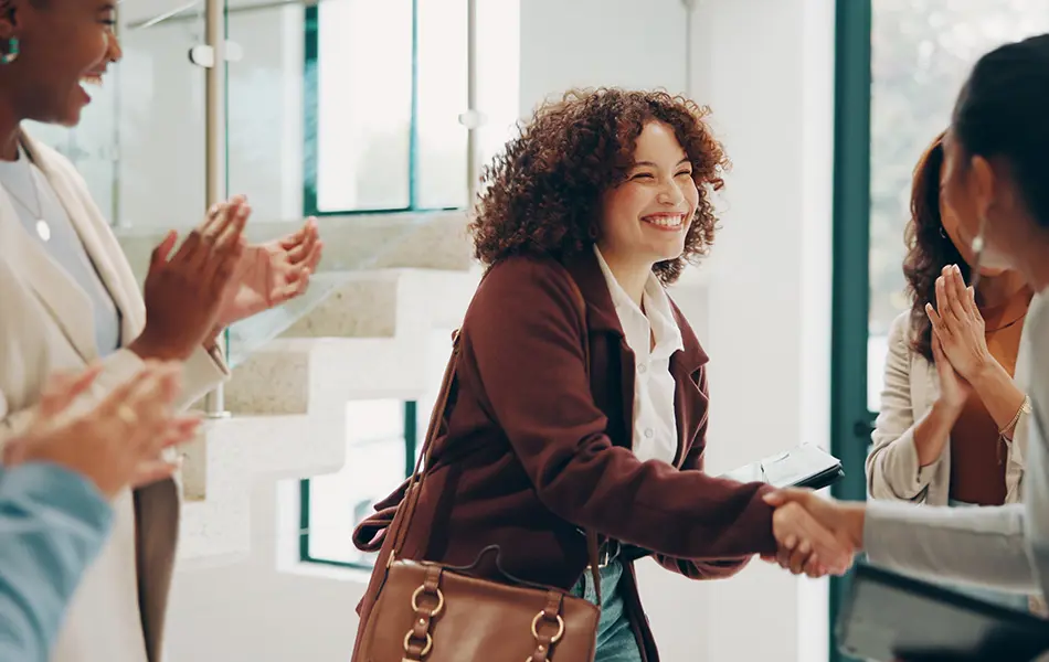 Smiling woman shaking hands while being welcomed by colleagues in a bright office space.
