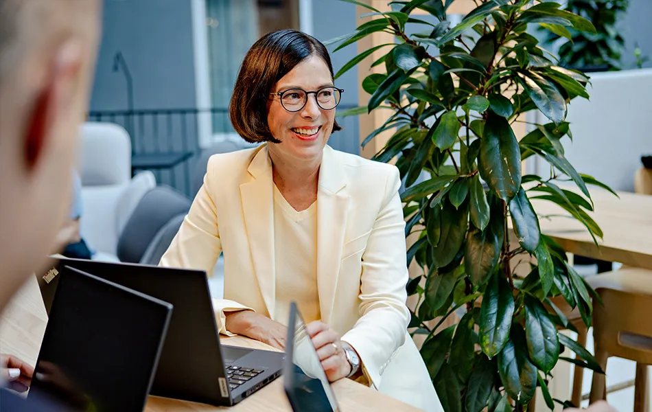 Leende kvinna i vit kavaj som sitter vid ett bord med bärbara datorer i ett modernt kontor.