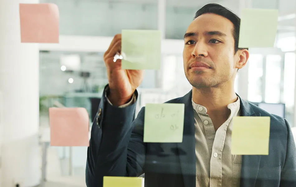 Fokuserad man som skriver på post-it-lappar fästa på en glasvägg i ett modernt kontor.