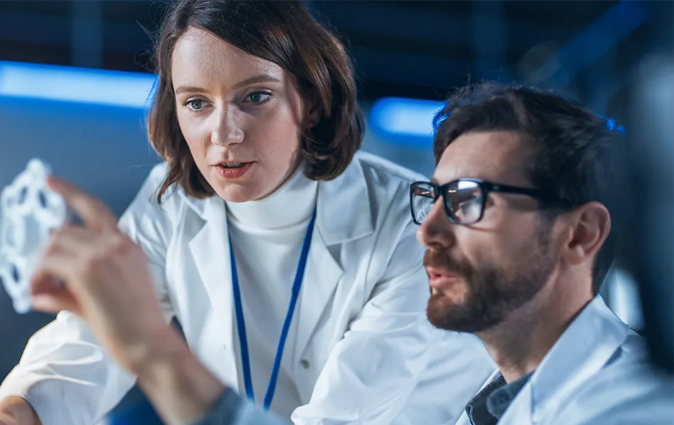 Två forskare i labbrockar analyserar ett 3D-utskrivet objekt i ett tekniskt laboratorium.